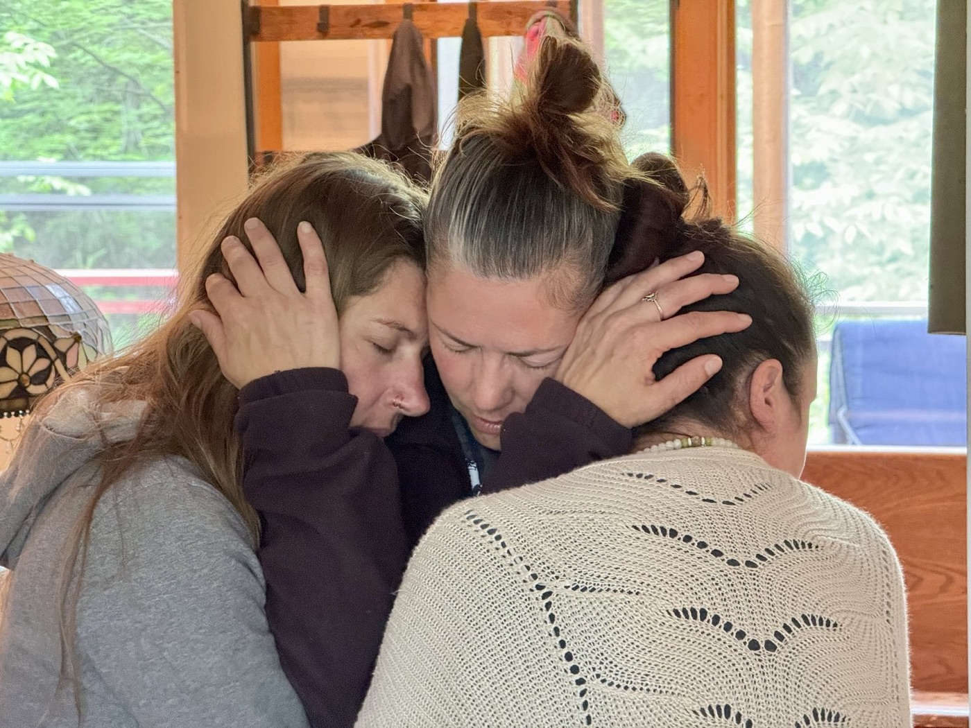 Three women holding each other in a close embrace by a window
